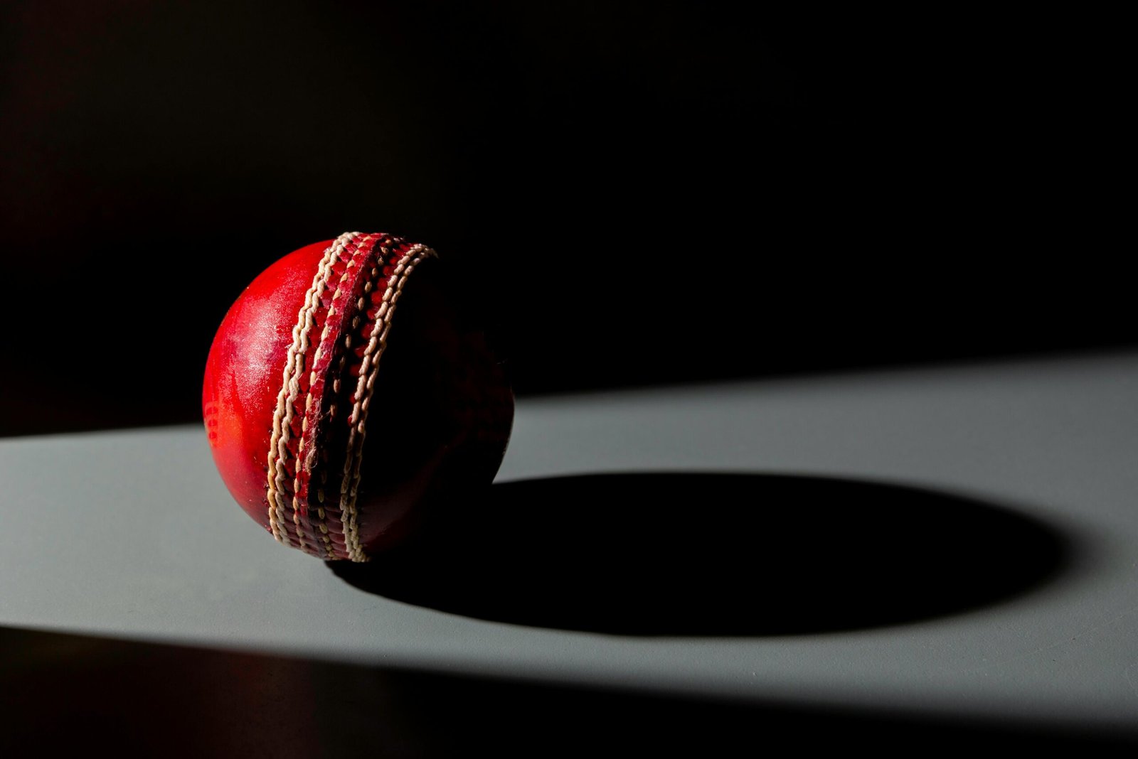 Dramatic image of a red cricket ball casting a shadow, highlighting texture and detail.